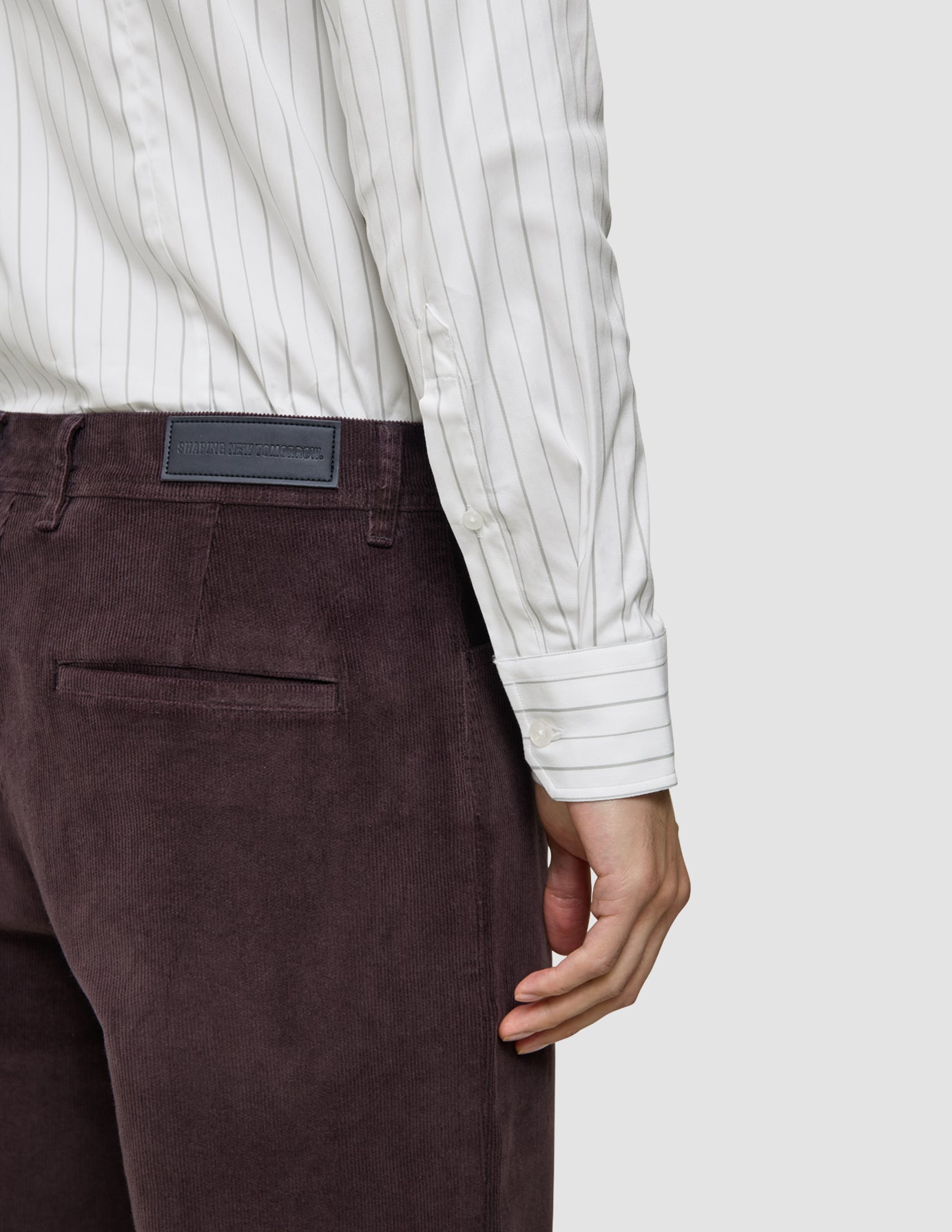 Ein hochgeschlagenes Hemd in Regular-Passform, hellgrau gestreift mit weißen, dünnen grauen Längsstreifen, in eine dunkelviolette Cordhose gesteckt, mit einem Markenetikett auf der Rückseite des Bunds.