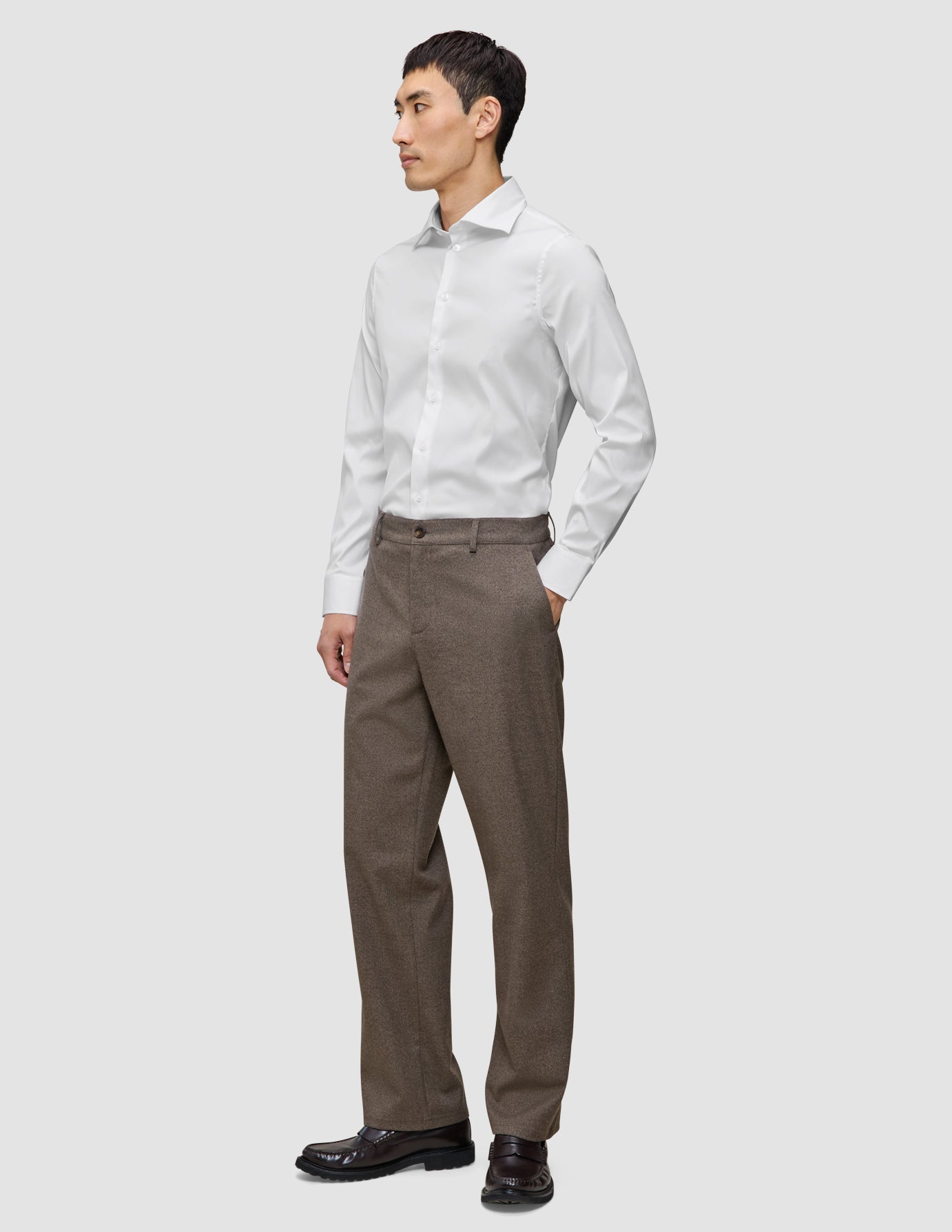 Ein Mann trägt ein weißes, schmal geschnittenes Elevated Shirt mit Knöpfen, das er in eine braune Hose gesteckt hat, sowie dunkle Slipper.