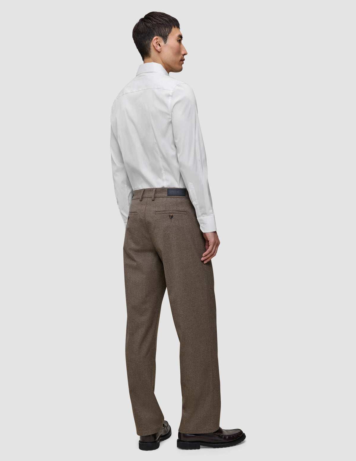 Ein Mann, von hinten gesehen, der ein weißes, schmal geschnittenes Elevated-Hemd trägt, das in eine braune Hose gesteckt ist, sowie dunkle Slipper.