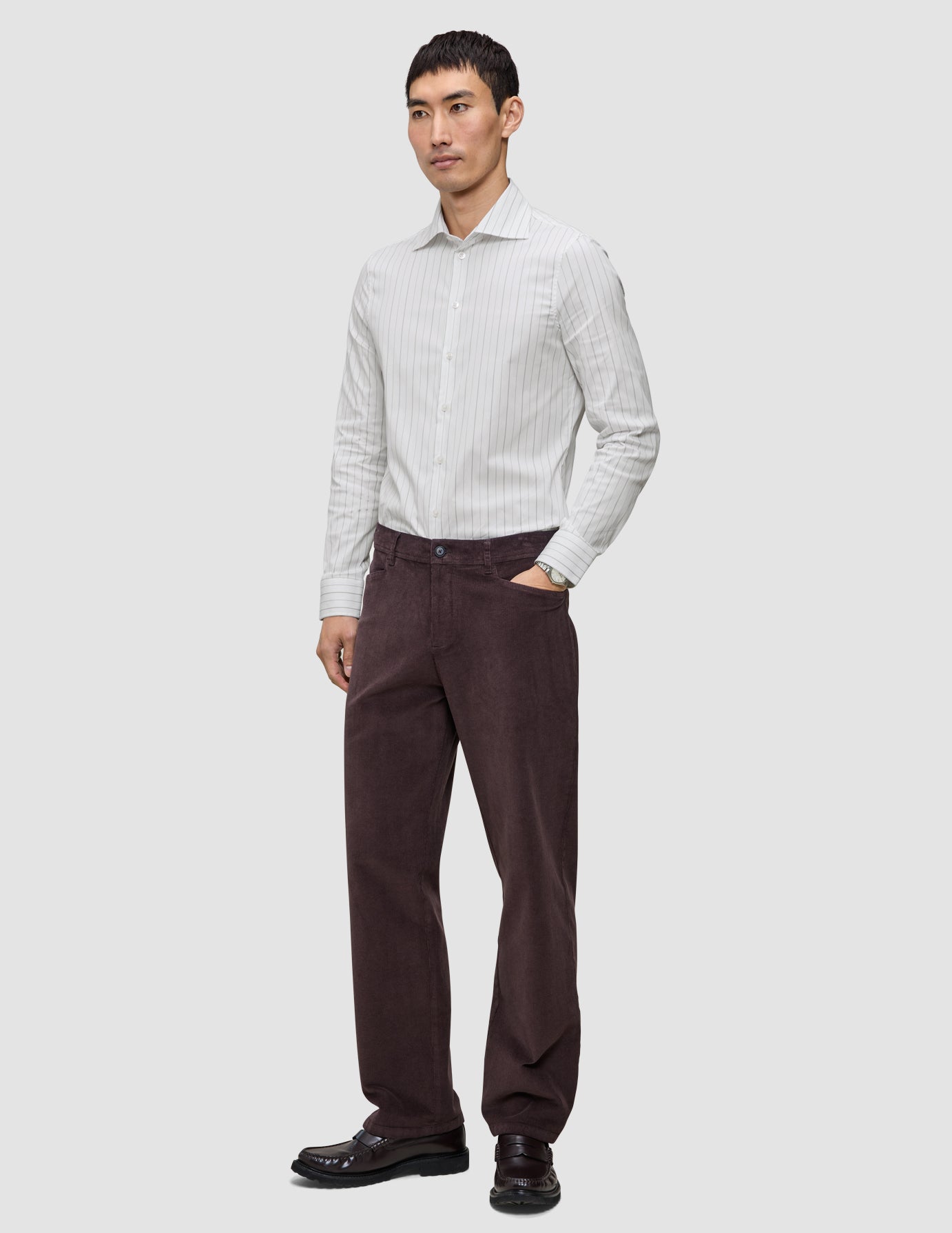 Ein Mann, der ein hellgrau gestreiftes Elevated Shirt Slim, eine dunkelbraune Hose und dunkle Slipper trägt.