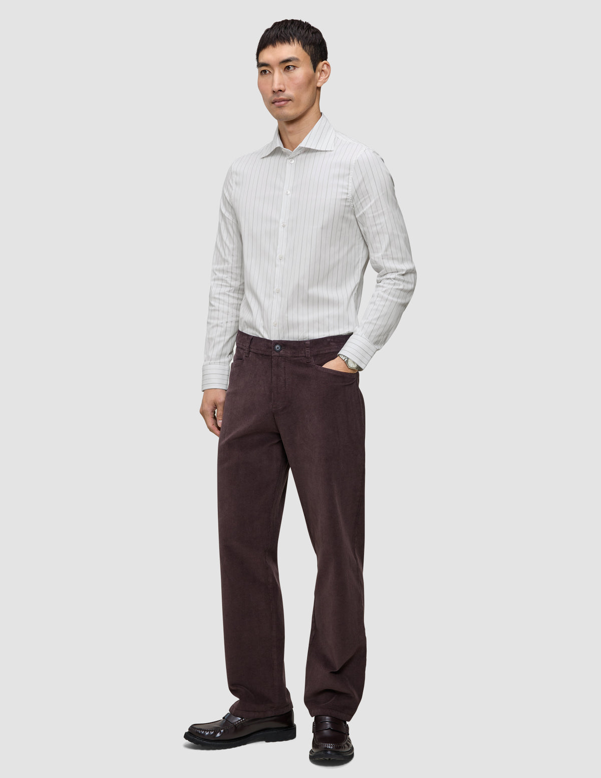 Ein Mann, der ein hellgrau gestreiftes Elevated Shirt Slim, eine dunkelbraune Hose und dunkle Slipper trägt.