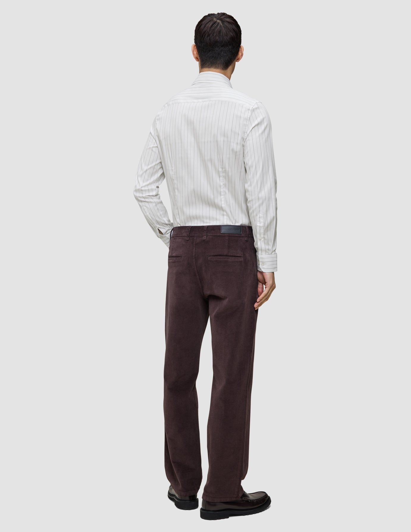 Eine Person, von hinten gesehen, die ein hellgrau gestreiftes Elevated Shirt Slim, eine dunkelbraune Hose und dunkle Slipper trägt.