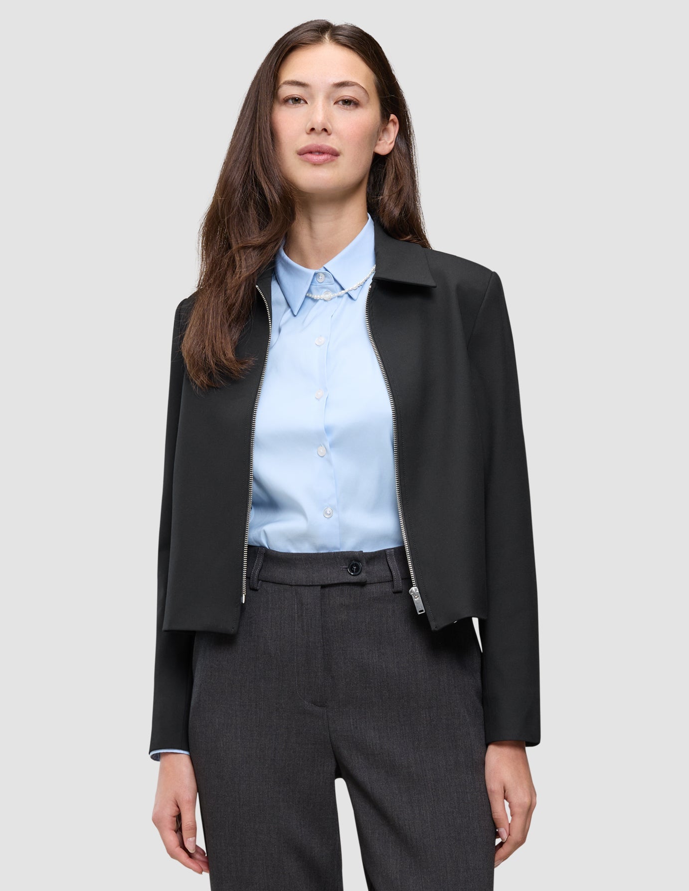 Eine Person, die eine schwarze Essential Short Zip Jacket über einem hellblauen Hemd mit Kragen und einer dunklen Hose trägt.