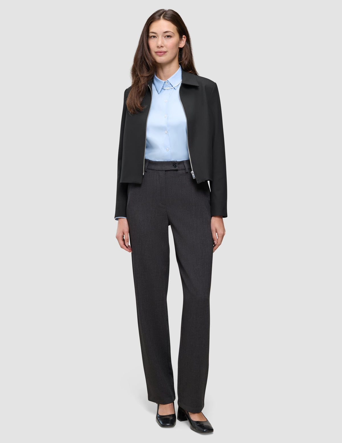 Eine Frau trägt eine schwarze Essential Short Zip Jacket über einem hellblauen Hemd und einer grauen Hose.