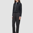Eine Person, die eine schwarze Essential Short Zip Jacket über einem hellblauen Hemd mit Kragen und einer dunkelgrauen Hose trägt; das Design besticht durch eine schlichte, skandinavische Ästhetik.