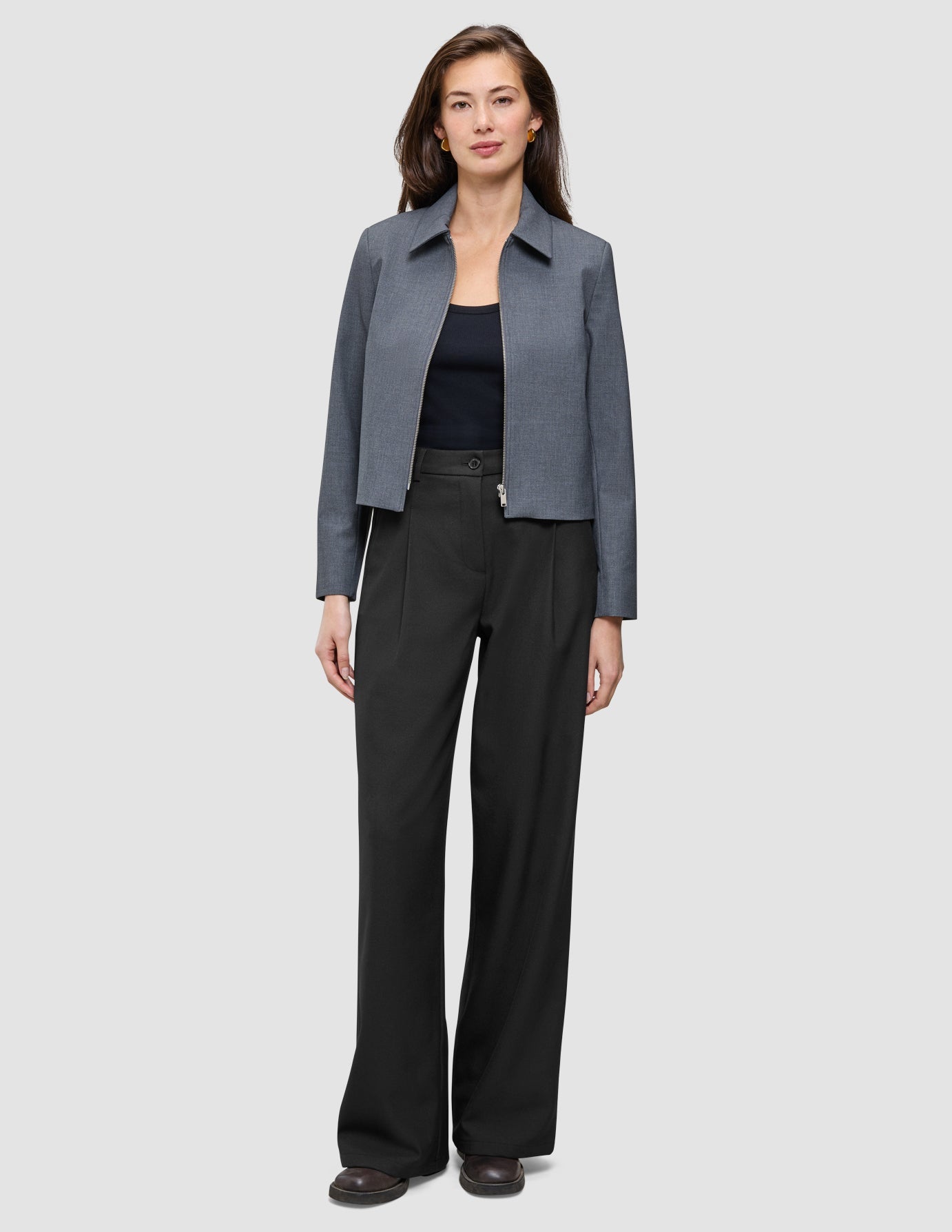 Eine Frau, die eine Essential Short Zip Jacket in Dunkelgrau-Melange über einem schwarzen Oberteil und einer schwarzen Hose mit weitem Bein trägt.