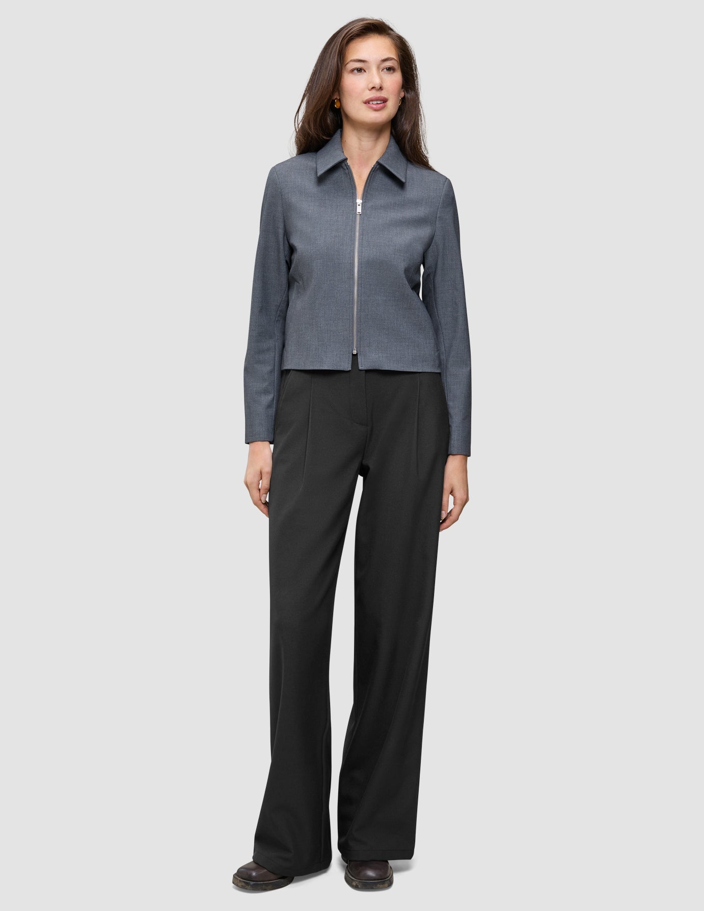 Eine Frau, die eine Essential Short Zip Jacket in Dunkelgrau-Melange mit Reißverschluss vorne und eine schwarze Hose mit weitem Bein trägt.