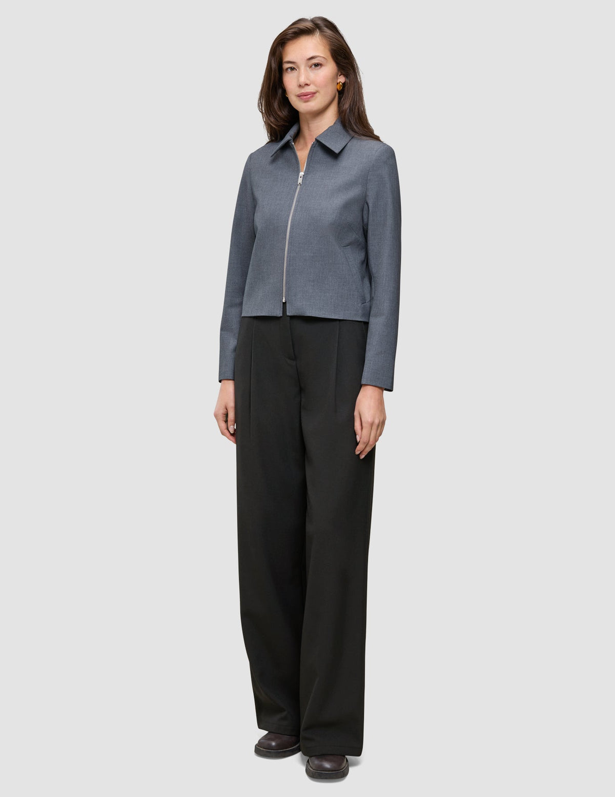 Eine Frau trägt eine Essential Short Zip Jacket in Dunkelgrau-Melange und eine schwarze Hose mit weitem Bein.