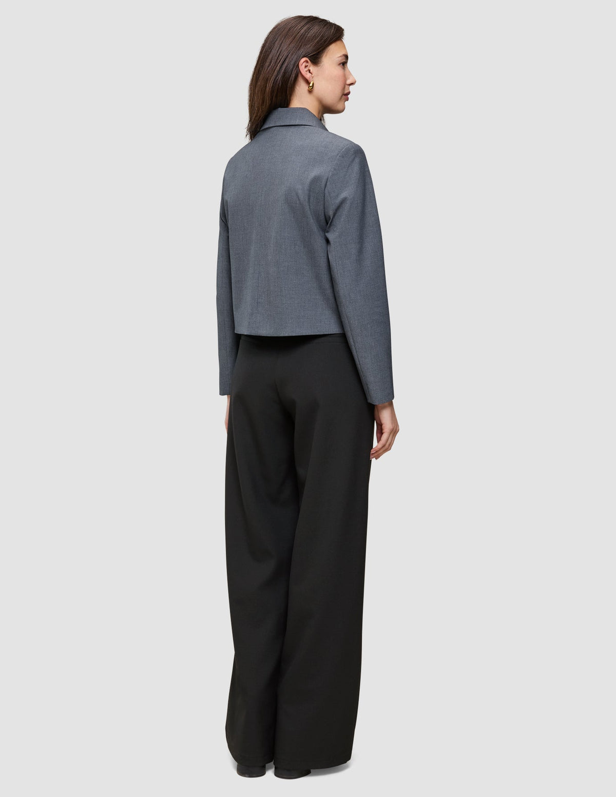 Eine Frau, von hinten gesehen, die eine Essential Short Zip Jacket in Dunkelgrau-Melange als kurze Reißverschlussjacke und eine schwarze Hose mit weitem Bein trägt.