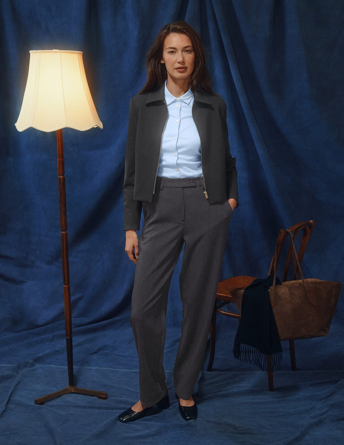 Eine Frau, die eine Essential Short Zip Jacket in Dunkelgrau-Melange über einem hellblauen Hemd und einer dunkelgrauen Hose trägt, was ein minimalistisches skandinavisches Design widerspiegelt.