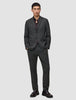 Ein Mann, der eine Jacke von Motion Blazer im Modell Regular in der Farbe Pepper Grey und eine dazu passende Hose über einem dunklen Hemd mit Knöpfen trägt, dazu dunkle Slipper in einem schlichten, skandinavischen Design.