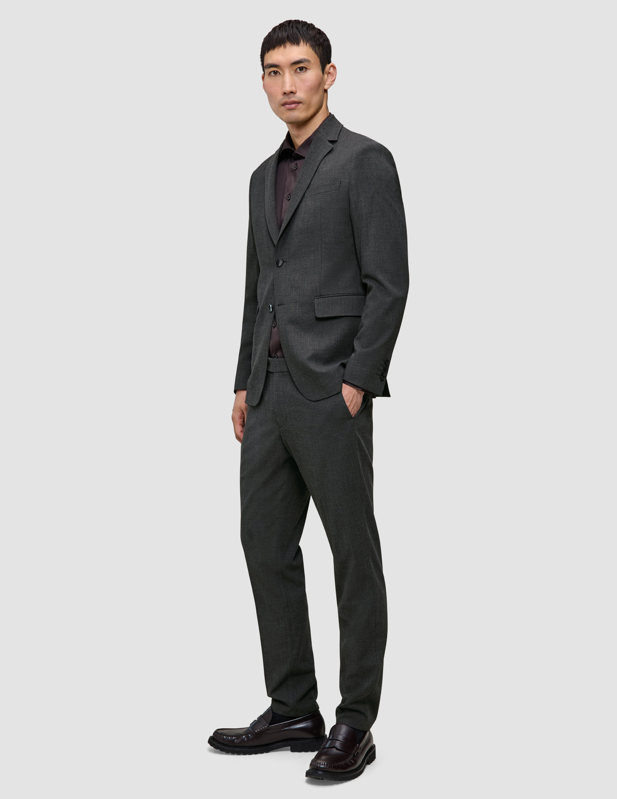 Ein Mann in einem Motion Blazer Regular in der Farbe Pepper Grey, einem dunklen Hemd mit Knopfleiste und dunklen Slippern steht da, die Hand in der Hosentasche.