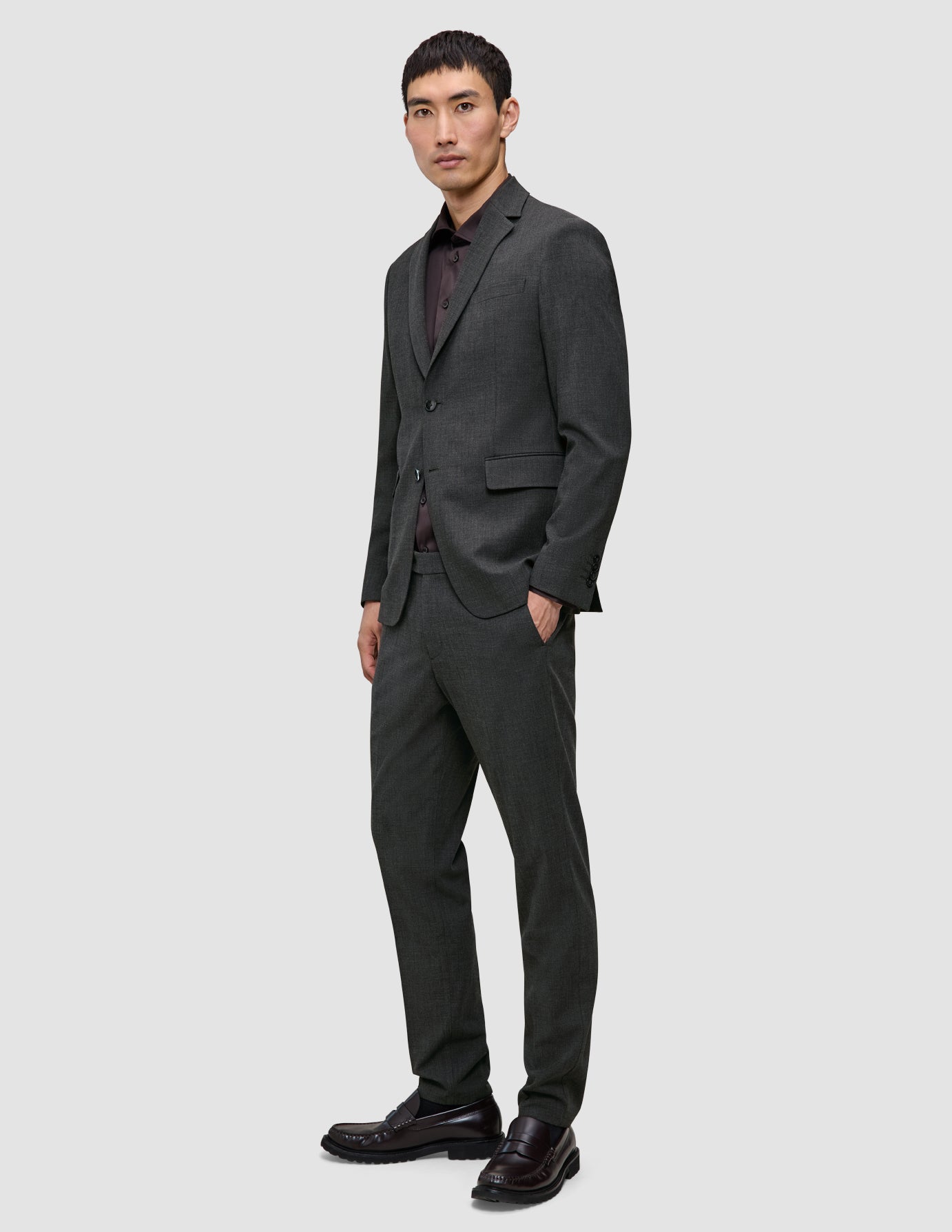 Ein Mann, der einen Motion Blazer Slim in der Farbe Pepper Grey mit einer dazu passenden Hose, einem dunklen Hemd und dunklen Loafern im klaren, skandinavischen Design trägt.