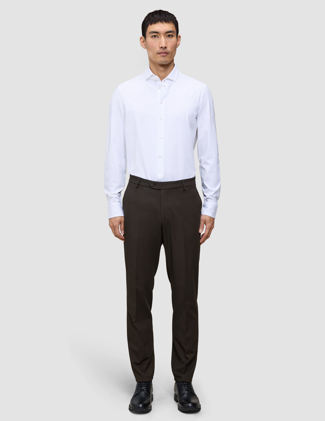 Ein Mann in einem weißen Hemd und einer Motion Suit-Hose in der Farbe Regular Cognac, kombiniert mit schwarzen Schuhen für einen eleganten Look.