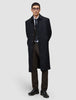 Ein Mann, der einen schwarzen Mantel über einem Anzug, einem weißen Hemd, einer Krawatte und einer Motion Suit Pants Regular Cognac trägt und vor einem hellgrauen Hintergrund steht, was ihm einen eleganten Look verleiht.