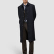 Ein Mann, der einen schwarzen Mantel über einem Anzug, einem weißen Hemd, einer Krawatte und einer Motion Suit Pants Regular Cognac trägt und vor einem hellgrauen Hintergrund steht, was ihm einen eleganten Look verleiht.