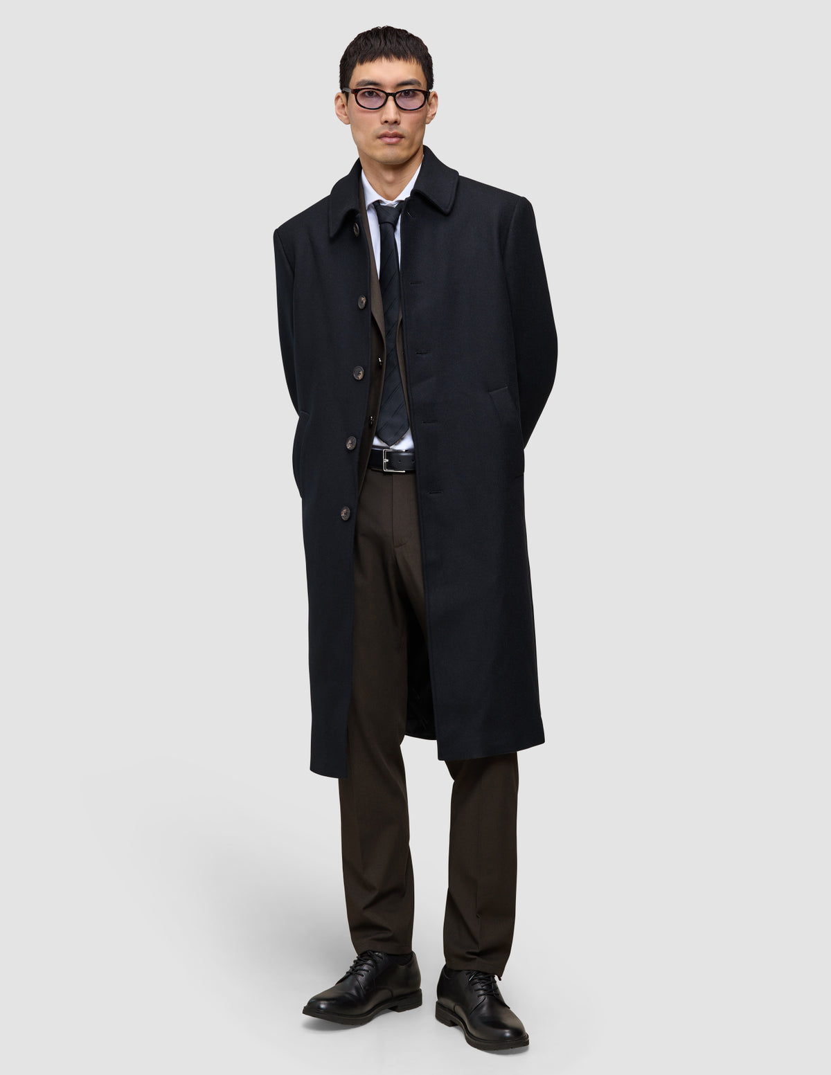 Ein Mann, der einen schwarzen Mantel über einem Anzug, einem weißen Hemd, einer Krawatte und einer Motion Suit Pants Regular Cognac trägt und vor einem hellgrauen Hintergrund steht, was ihm einen eleganten Look verleiht.