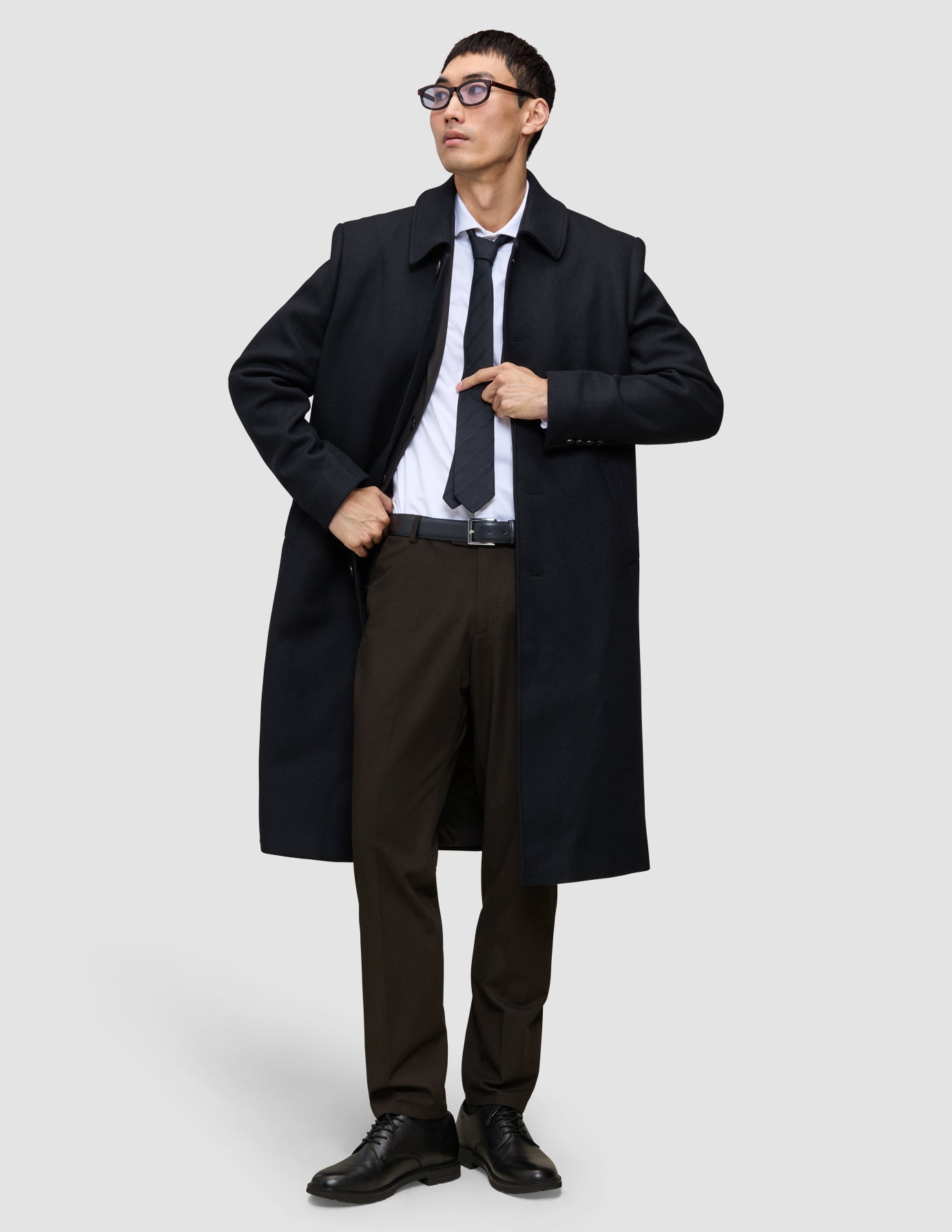 Ein Mann trägt einen schwarzen Mantel, ein weißes Hemd und eine dunkle Krawatte sowie die Motion Suit Pants Regular Cognac – eine elegante, braune Anzughose mit 360°-Stretch.