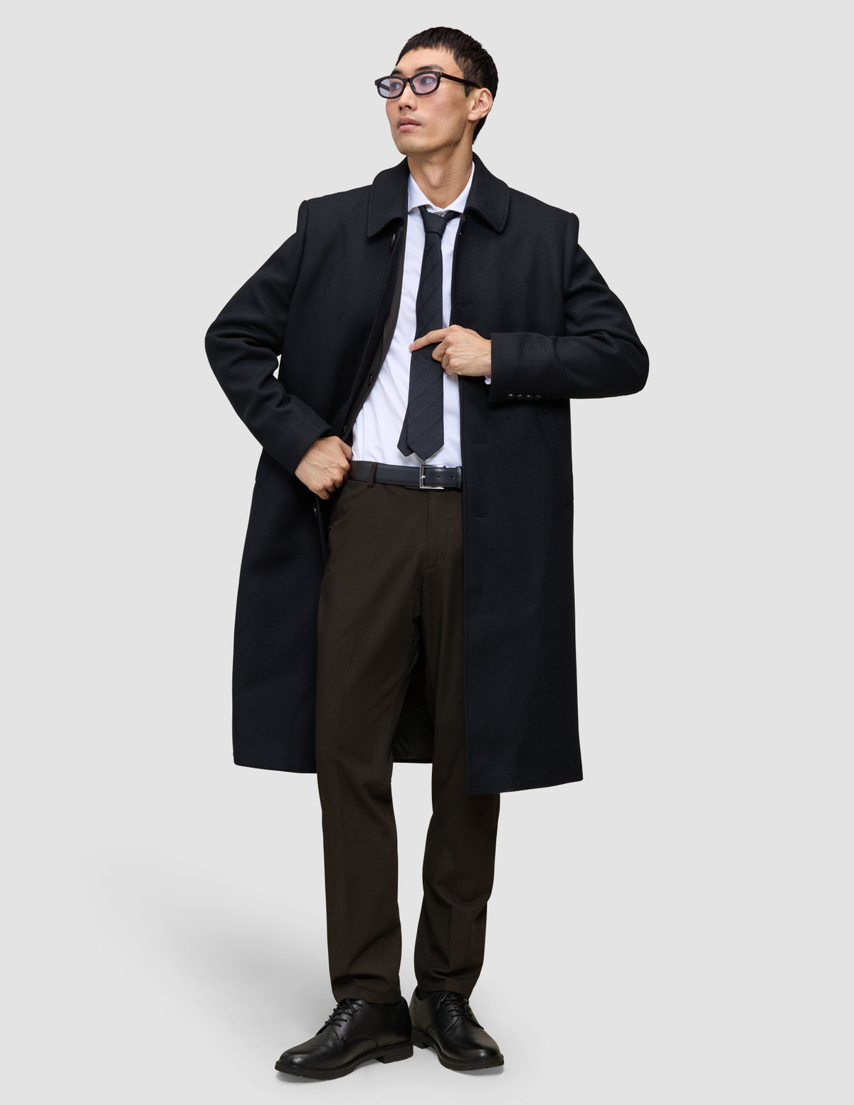 Ein Mann trägt einen schwarzen Mantel, ein weißes Hemd und eine dunkle Krawatte sowie die Motion Suit Pants Regular Cognac – eine elegante, braune Anzughose mit 360°-Stretch.