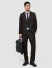 Ein Mann in einer eleganten dunkelbraunen Motion-Anzughose Regular Cognac, einem weißen Hemd und einer schwarzen Krawatte hält einen schwarzen Rucksack in der Hand.