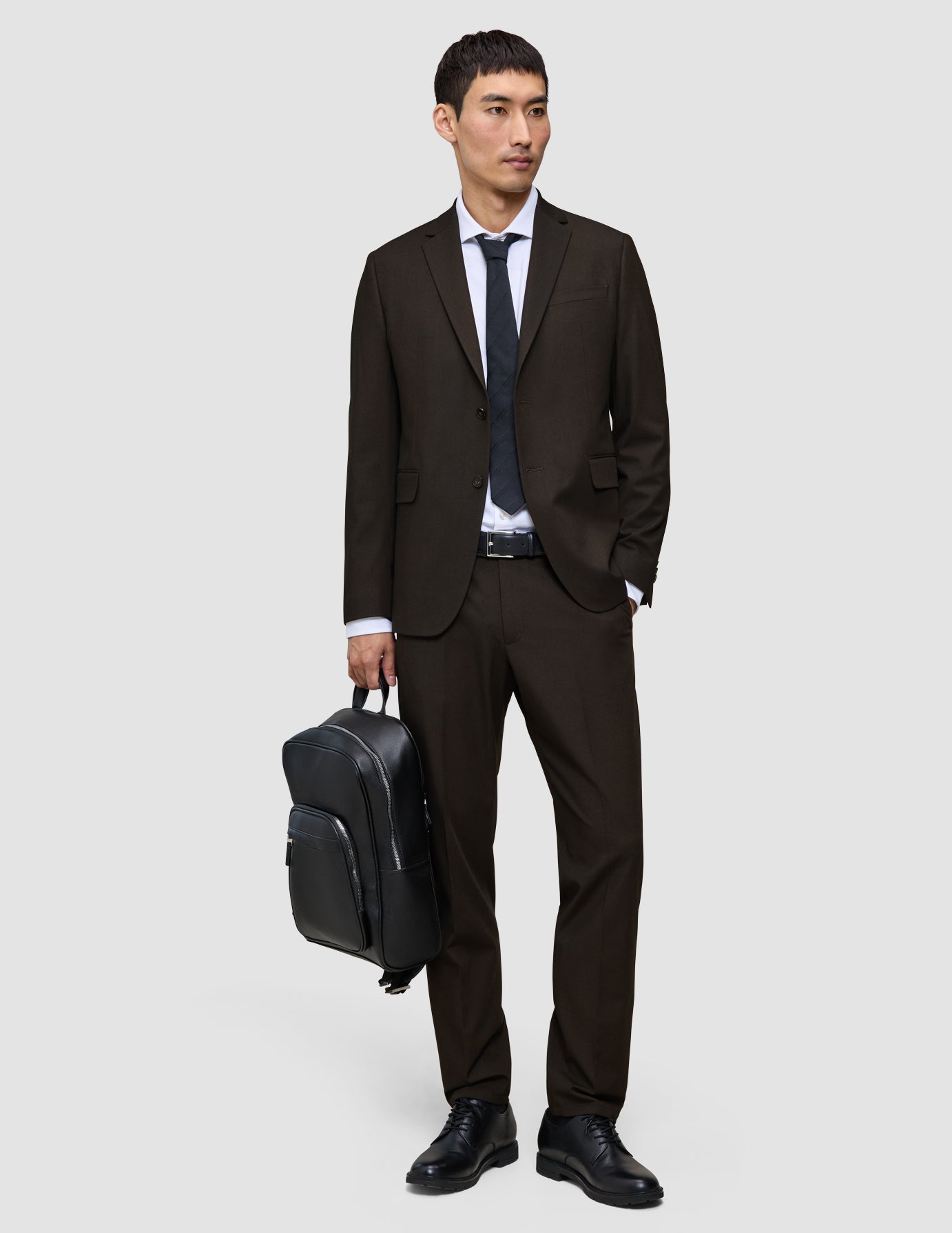 Ein Mann in einer eleganten dunkelbraunen Motion-Anzughose Regular Cognac, einem weißen Hemd und einer schwarzen Krawatte hält einen schwarzen Rucksack in der Hand.