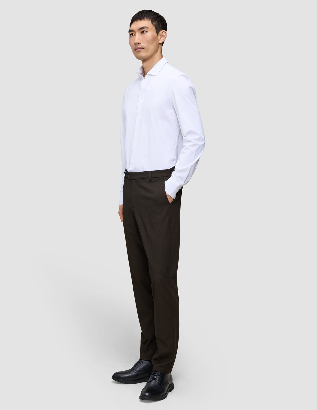Ein Mann, im Profil zu sehen, der ein weißes, langärmeliges Hemd mit Knöpfen, eine Motion Suit-Hose in der Passform Regular in der Farbe Cognac sowie schwarze Anzugschuhe trägt und damit einen eleganten Look präsentiert.
