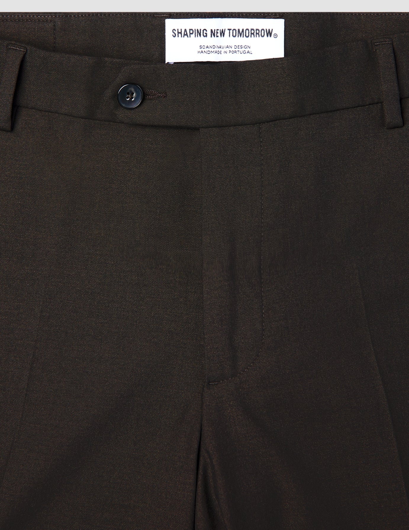 Nahaufnahme einer braunen Motion Suit-Hose in der Passform Regular in der Farbe Cognac mit Knopfverschluss und einem Etikett am Bund mit der Aufschrift SHAPING NEW TOMORROW, SCANDINAVIAN DESIGN, HANDMADE IN PORTUGAL.