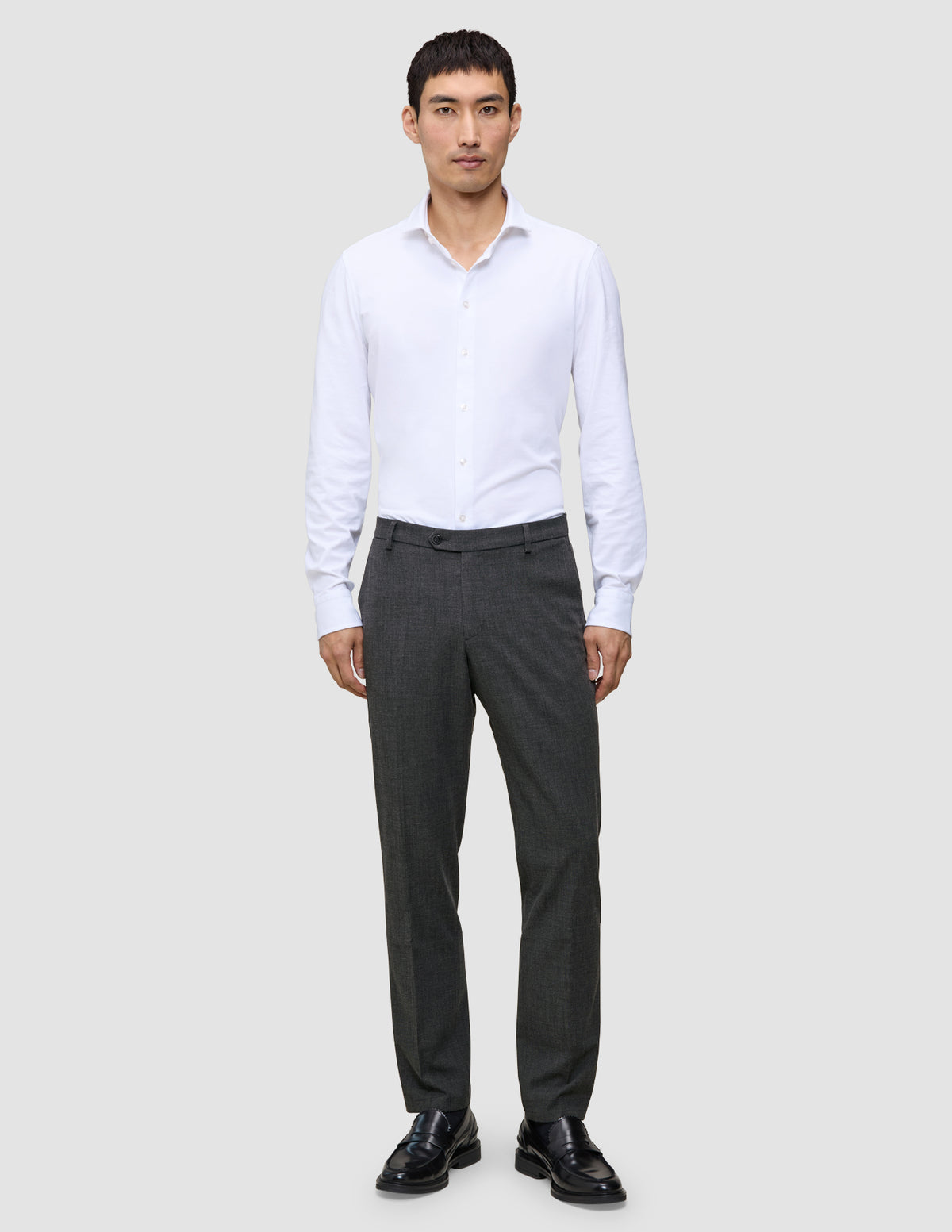 Ein Mann, der ein weißes Hemd, eine Motion Suit-Hose in der Passform Regular in der Farbe Pepper Grey und schwarze Anzugschuhe trägt.