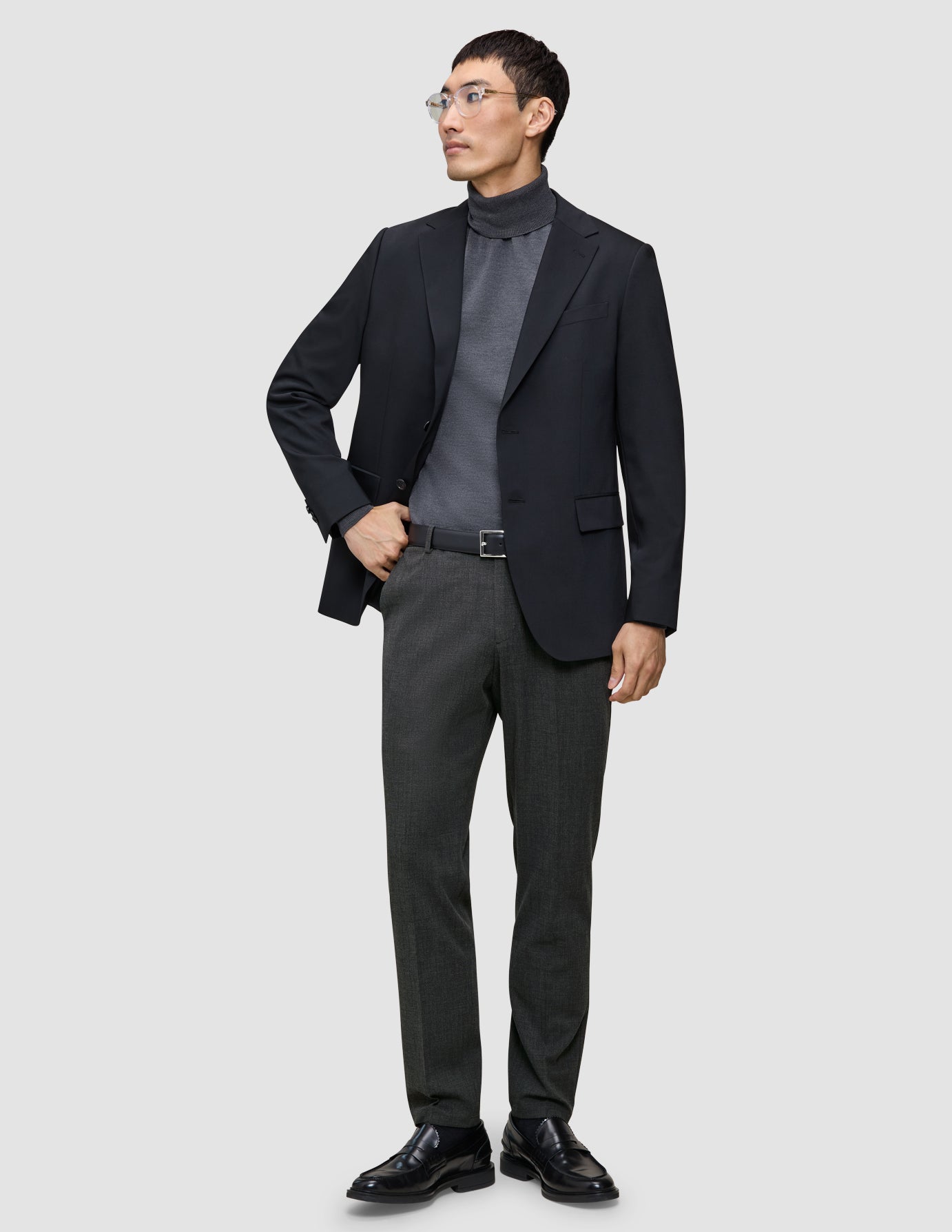 Ein Mann, der einen schwarzen Blazer, einen grauen Rollkragenpullover und eine Motion Suit-Hose in der Farbe Regular Pepper Grey trägt, steht da, die Hand in der Tasche.