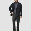 Ein Mann, der einen schwarzen Blazer, einen grauen Rollkragenpullover und eine Motion Suit-Hose in der Farbe Regular Pepper Grey trägt, steht da, die Hand in der Tasche.