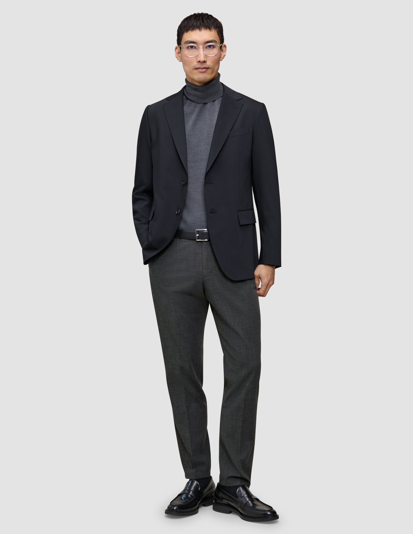 Ein Mann, der einen schwarzen Blazer, einen grauen Rollkragenpullover, eine Motion Suit-Hose in der Passform Regular in der Farbe Pepper Grey sowie schwarze Slipper trägt.