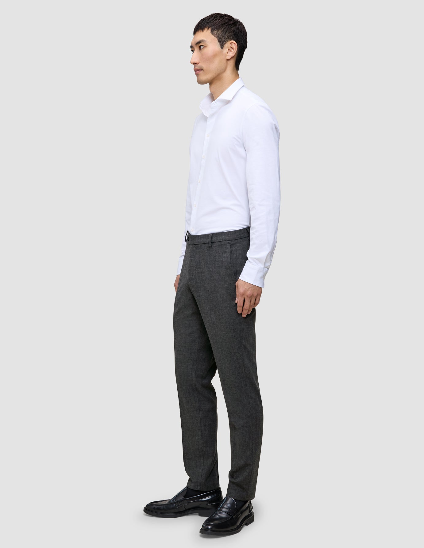 Ein Mann in einem weißen Hemd und einer grauen Motion Suit-Hose in der Farbe Regular Pepper Grey steht im Profil und trägt schwarze Slipper.
