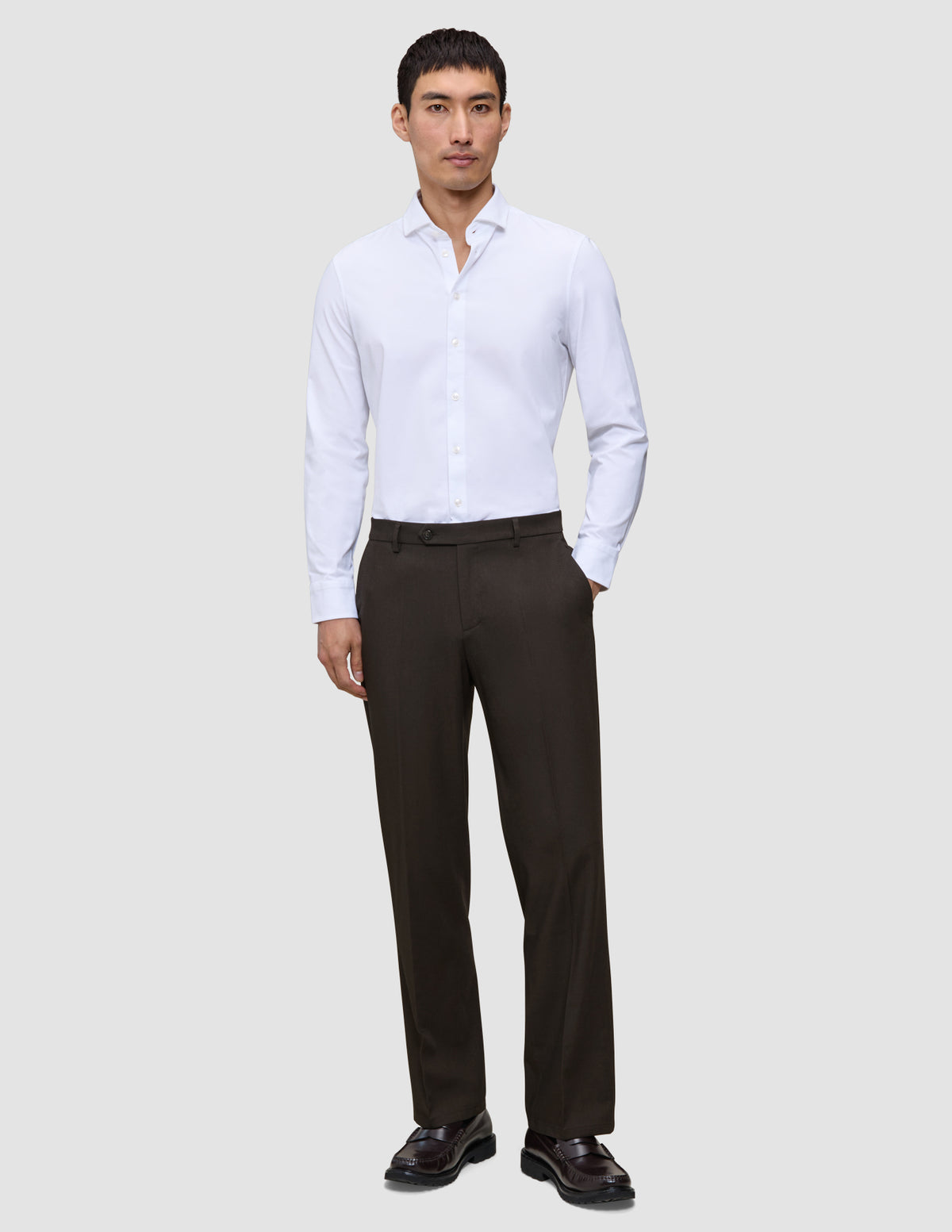 Ein Mann, der ein weißes, langärmeliges Hemd mit Knöpfen und eine Motion Suit Pants Relaxed Cognac sowie dunkle Loafer trägt.