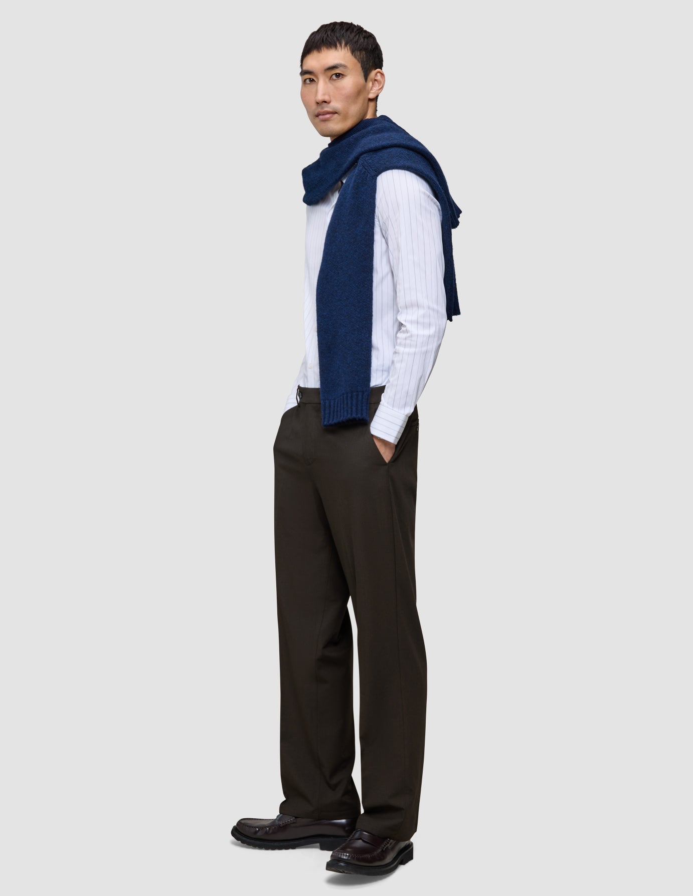 Ein Mann trägt ein weißes, fein gestreiftes Hemd mit Knöpfen, einen marineblauen Pullover, der über seinen Schultern liegt, sowie die Motion Suit Pants Relaxed Cognac – eine dunkelbraune Anzughose mit 360°-Stretch und lockerer Passform.
