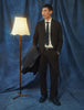 Ein Mann in einem Motion Suit Pants Relaxed Cognac-Anzug, dazu ein weißes Hemd und eine Krawatte, steht neben einer Stehlampe.