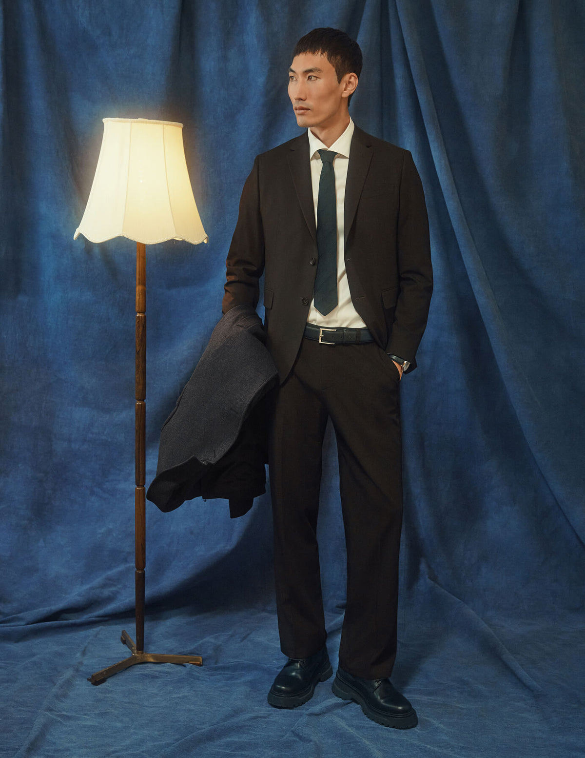 Ein Mann in einem Motion Suit Pants Relaxed Cognac-Anzug, dazu ein weißes Hemd und eine Krawatte, steht neben einer Stehlampe.