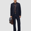 Ein Mann, der eine Motion Suit Pants Relaxed in Mitternachtsblau, ein dunkles Hemd und Slipper trägt und eine braune Lederaktentasche in der Hand hält.