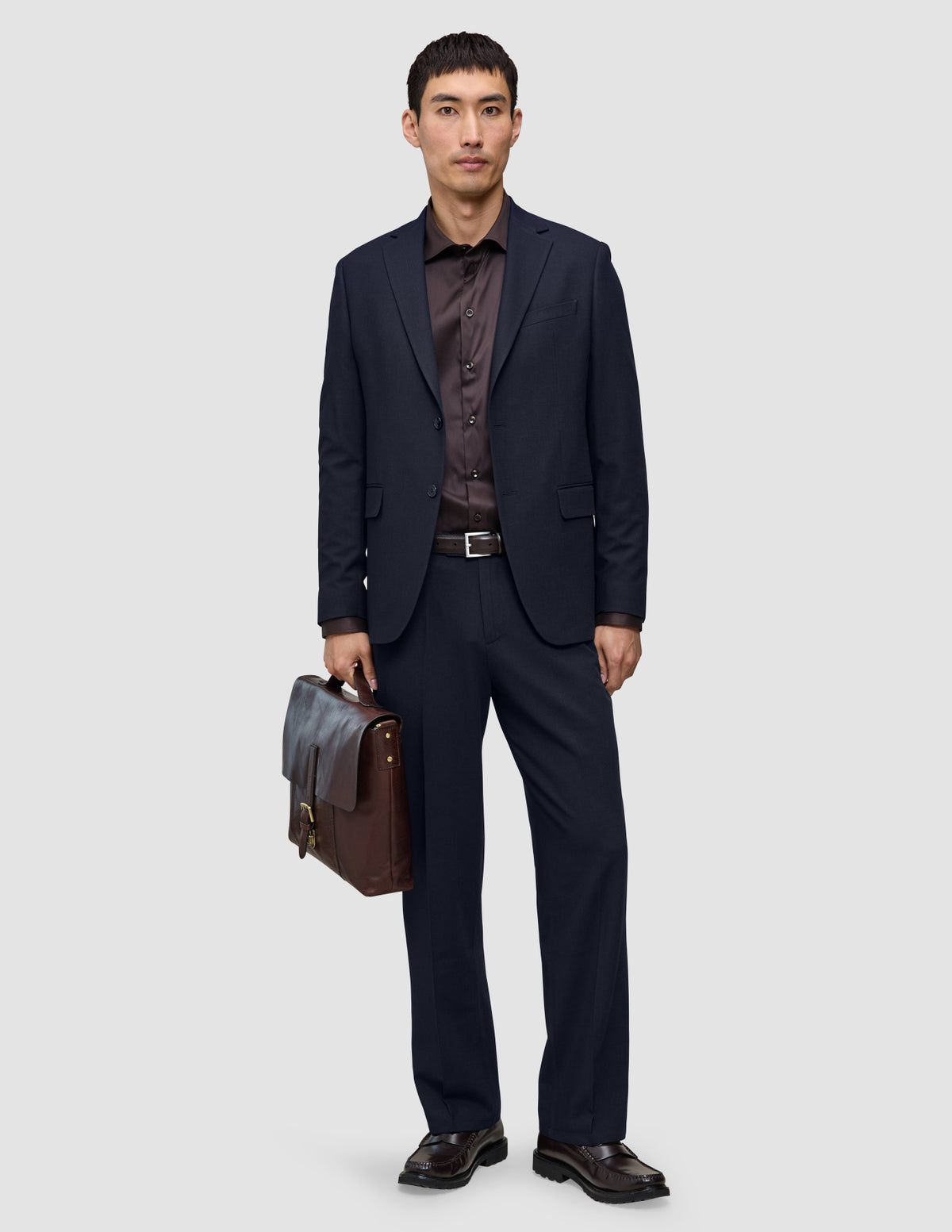 Ein Mann, der eine Motion Suit Pants Relaxed in Mitternachtsblau, ein dunkles Hemd und Slipper trägt und eine braune Lederaktentasche in der Hand hält.