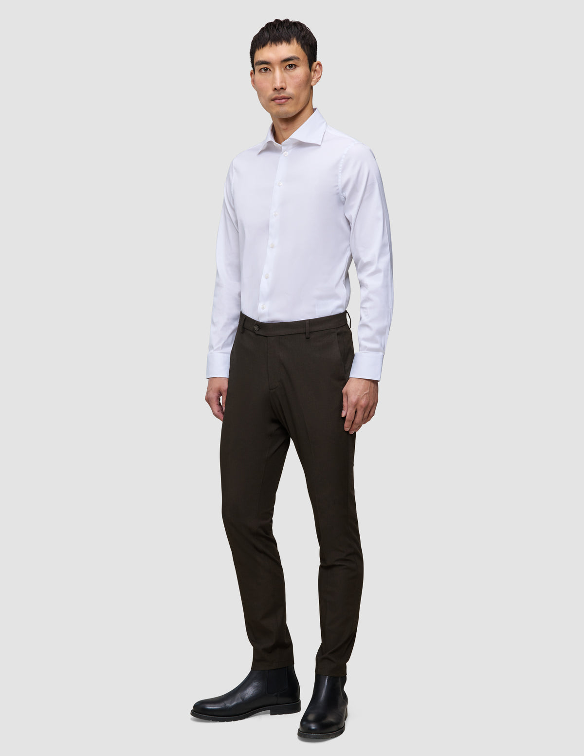 Ein Mann in einem weißen Hemd und einer schmal geschnittenen Motion Suit-Hose in der Farbe Slim Cognac, kombiniert mit schwarzen Lederstiefeln für einen eleganten Look.