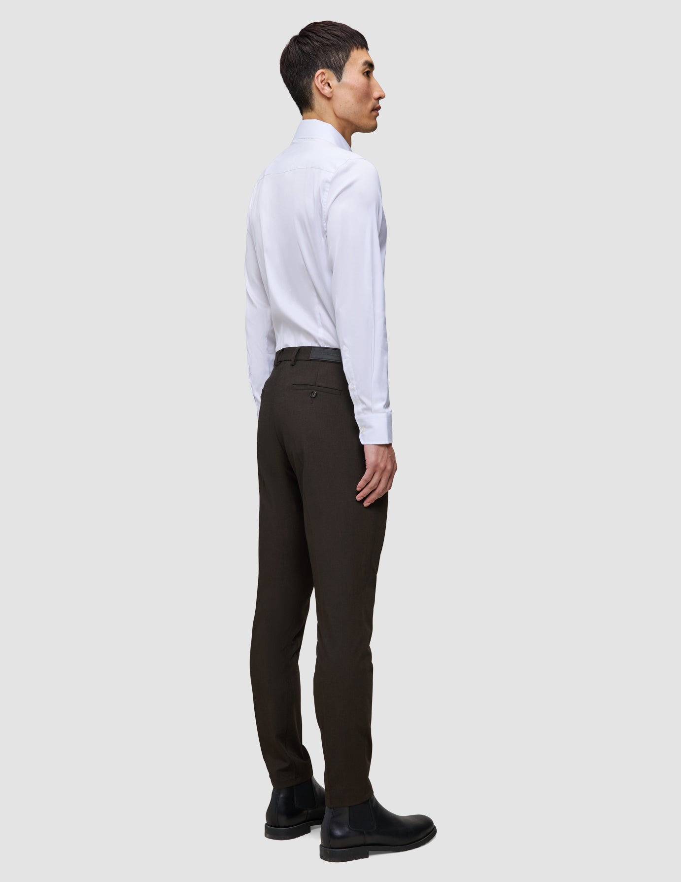 Ein Mann steht im Profil da und trägt ein weißes Hemd sowie eine Motion Suit Pants Slim Cognac – ein eleganter Look.
