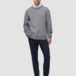 Ein Mann, der einen grauen Pullover mit Rundhalsausschnitt über einem weißen Hemd mit Kragen trägt, dazu eine Motion-Anzughose in der Farbe Slim Midnight Blue und braune Slipper.