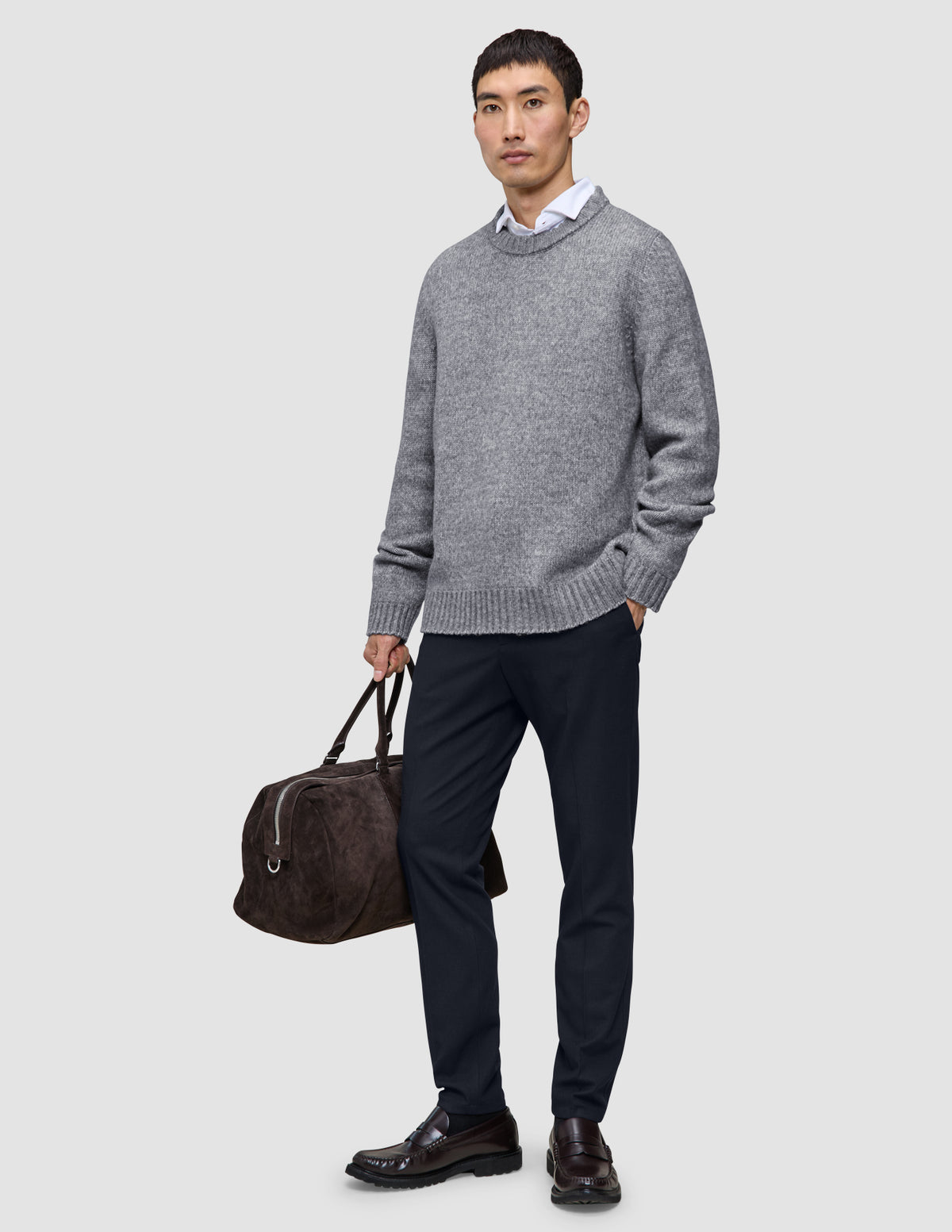 Ein Mann, der einen grauen Pullover über einem weißen Hemd mit Kragen und eine Motion Suit-Hose in der Farbe Slim Midnight Blue trägt und eine braune Reisetasche bei sich hat.