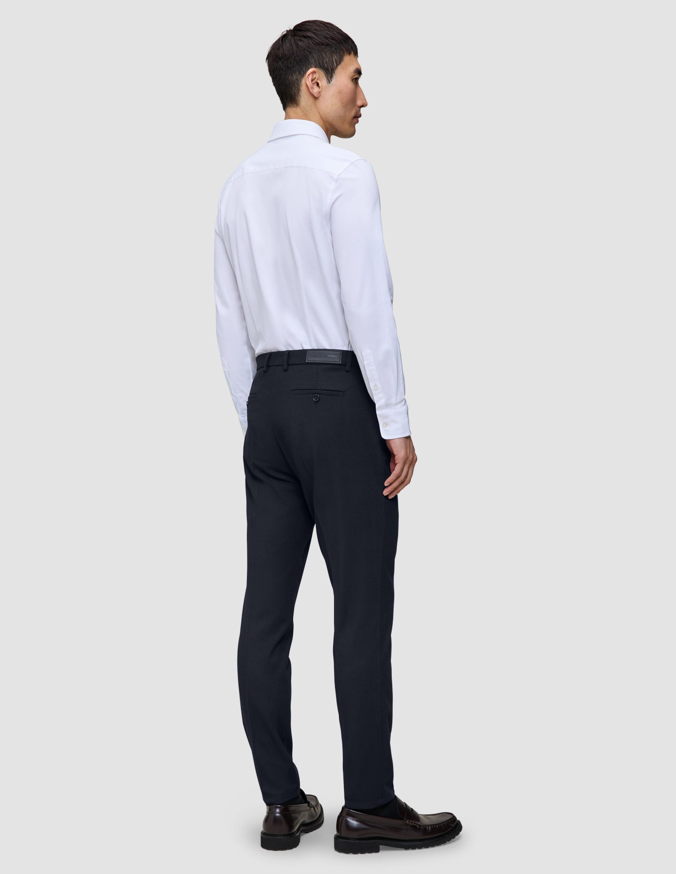 Eine Person, von hinten gesehen, die ein weißes Hemd und eine Motion Suit Pants Slim in Mitternachtsblau sowie dunkle Loafer trägt.