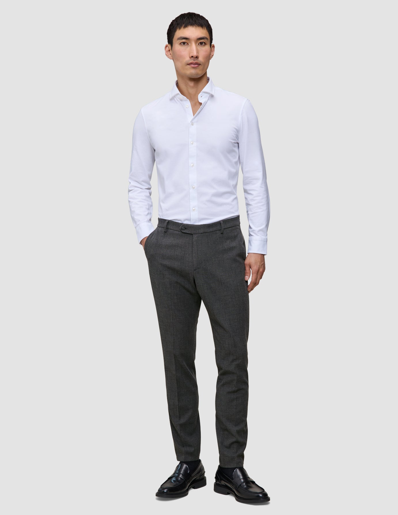Ein Mann, der ein weißes Hemd, eine Motion Suit-Hose in der Farbe Slim Pepper Grey und schwarze Slipper trägt.