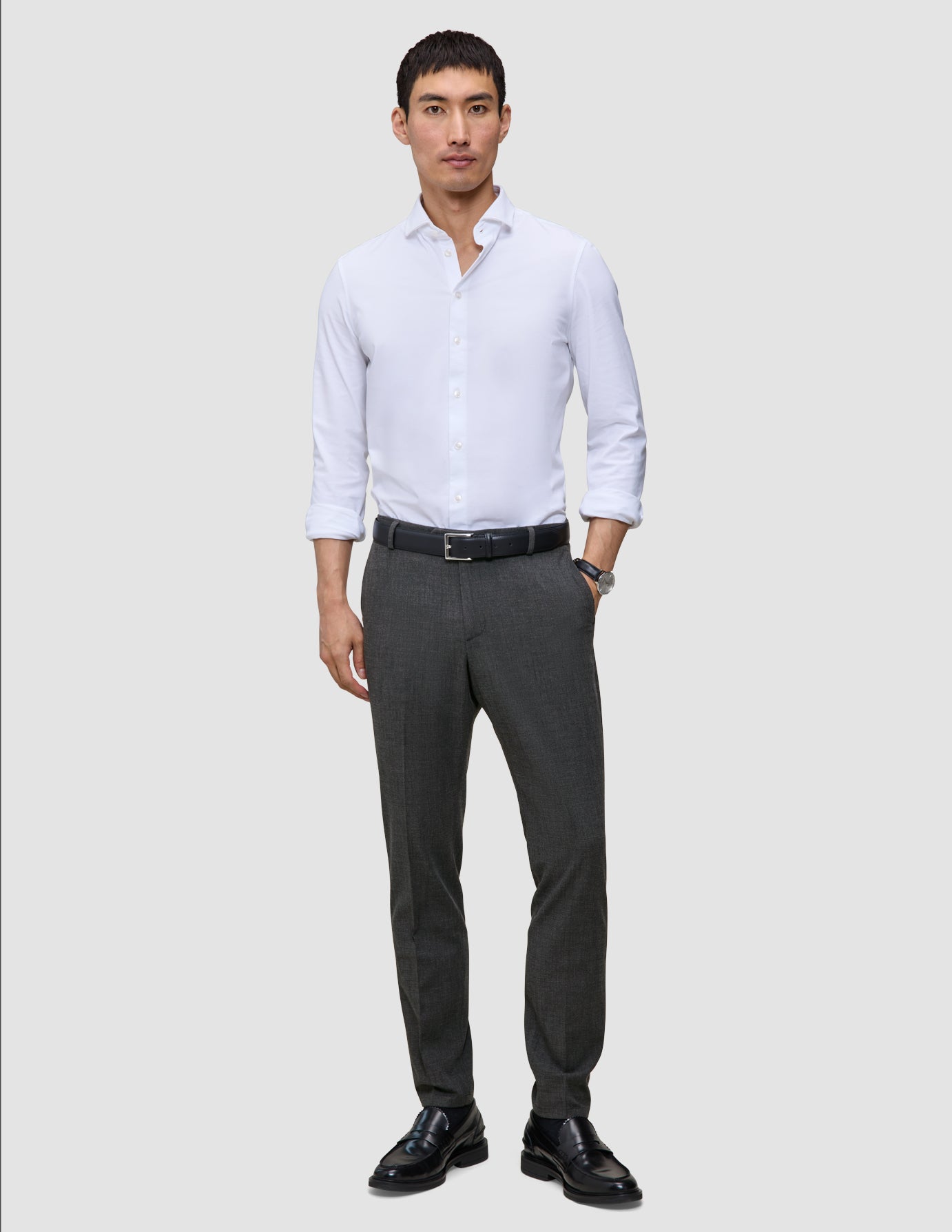 Ein Mann, der ein weißes Hemd, einen schwarzen Gürtel, eine Motion Suit-Hose in der Farbe Slim Pepper Grey und schwarze Slipper trägt.