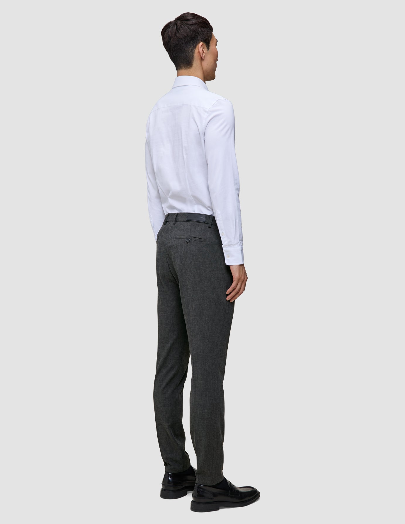 Ein Mann, von hinten gesehen, der ein weißes Hemd, eine Motion Suit-Hose in der Farbe Slim Pepper Grey und schwarze Slipper trägt.