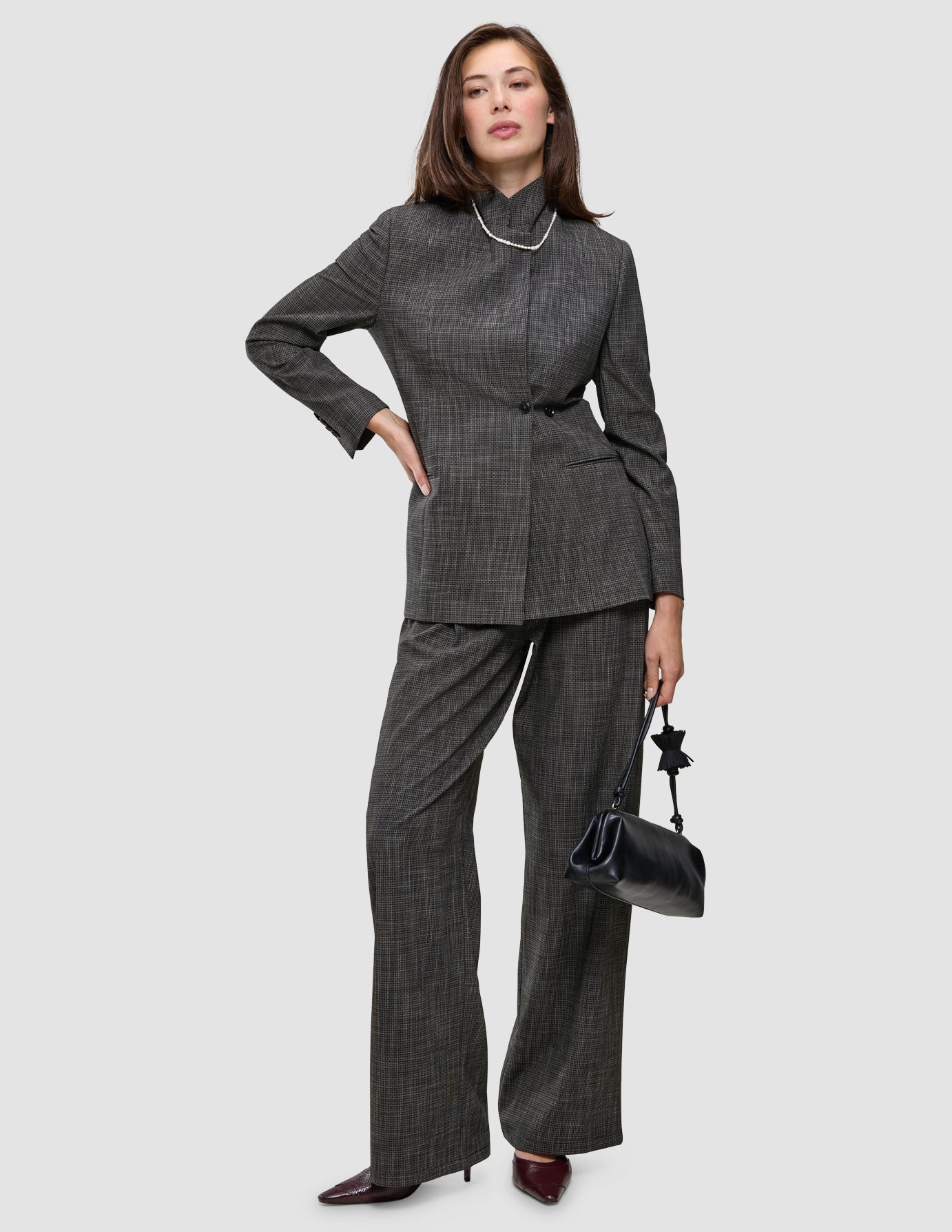 Eine Frau trägt ein auffälliges Kostüm in anthrazitgrauem Karomuster, bestehend aus einem Blazer und einer Hose mit weitem Bein, dazu eine Perlenkette und eine schwarze Handtasche.