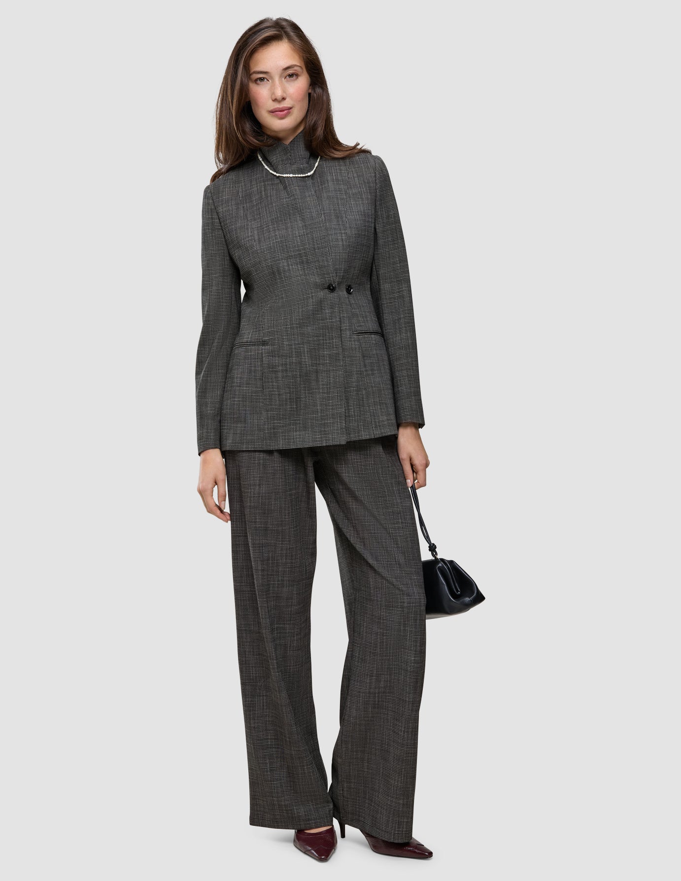 Eine Frau trägt das Outfit in Anthrazit, bestehend aus einem zweireihigen Nara-Blazer und einer dazu passenden Nara-Hose mit weitem Bein, ergänzt durch eine Perlenkette und eine kleine schwarze Handtasche.