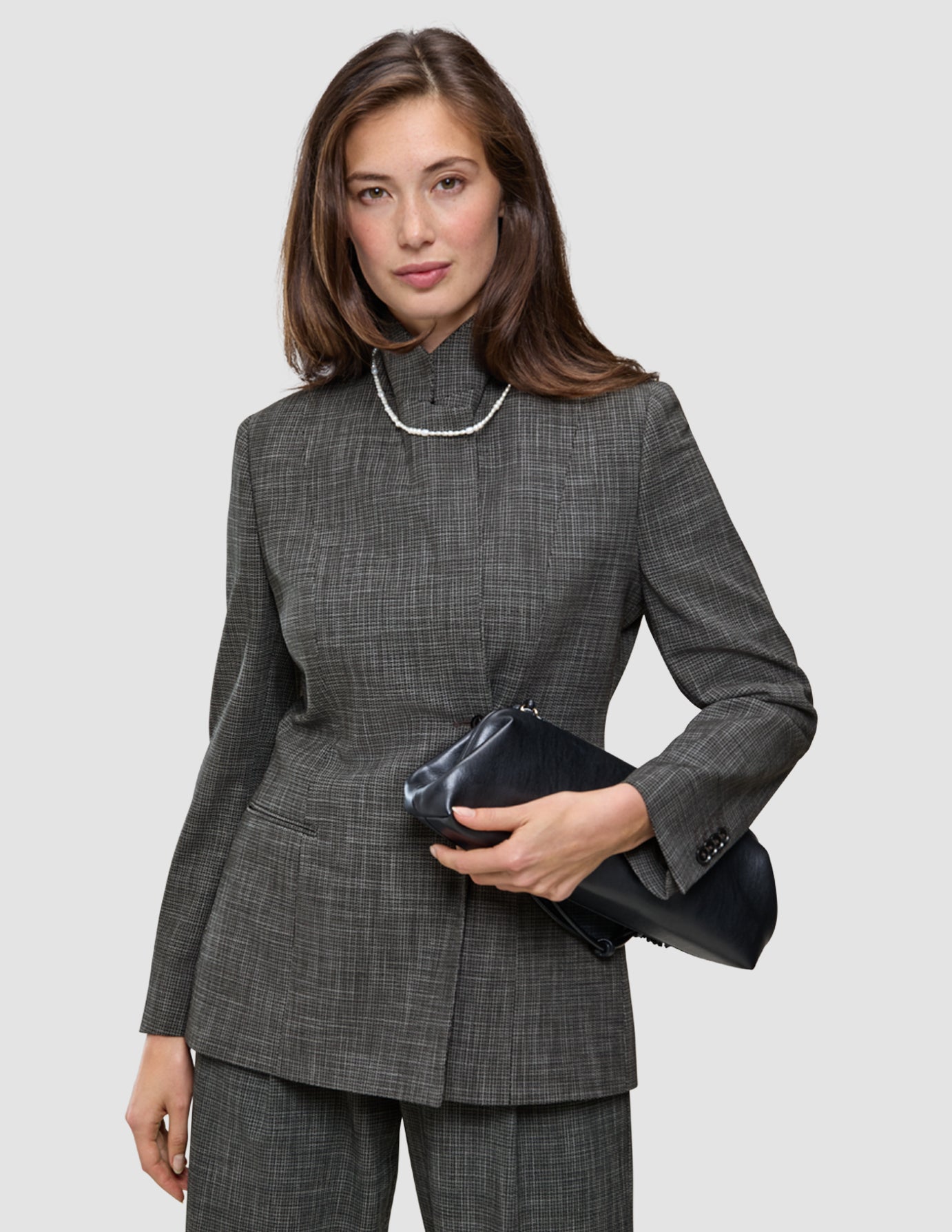 Eine Frau, die den Anzug trägt – ein grauer, karierter Nara-Blazer mit hohem Kragen und dazu passende Nara-Hose –, hält eine schwarze Clutch in der Hand.