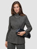 Eine Frau, die den Anzug trägt – ein grauer, karierter Nara-Blazer mit hohem Kragen und dazu passende Nara-Hose –, hält eine schwarze Clutch in der Hand.